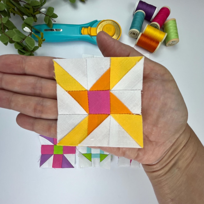 Peut inclure: Un bloc de courtepointe en tissu carr&eacute; avec un carr&eacute; rose central entour&eacute; de triangles orange et jaunes et de pi&egrave;ces de tissu blanc. Le bloc est tenu dans une paume, avec un cutter rotatif turquoise et plusieurs bobines de fil color&eacute;es.