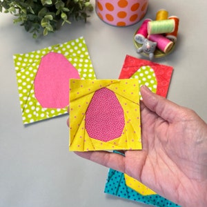 May include: A hand holding a small, square fabric block with a yellow polka dot border and a pink egg-shaped center. Other blocks with similar designs in green, red, and blue are visible, along with spools of thread and a polka dot container.