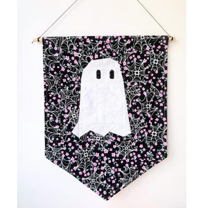 May include: A fabric wall hanging featuring a white ghost appliqué centered on a black background. The fabric is patterned with white spiderwebs and small pink flowers. The banner is suspended from a wooden dowel with a thin black string.