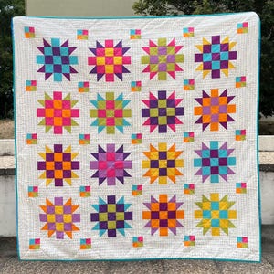 Pode incluir: Um edredom branco com uma borda turquesa que apresenta um padrão repetitivo de blocos em forma de estrela coloridos. Cada bloco é composto por quadrados menores em tons de roxo, laranja, amarelo, verde e azul.