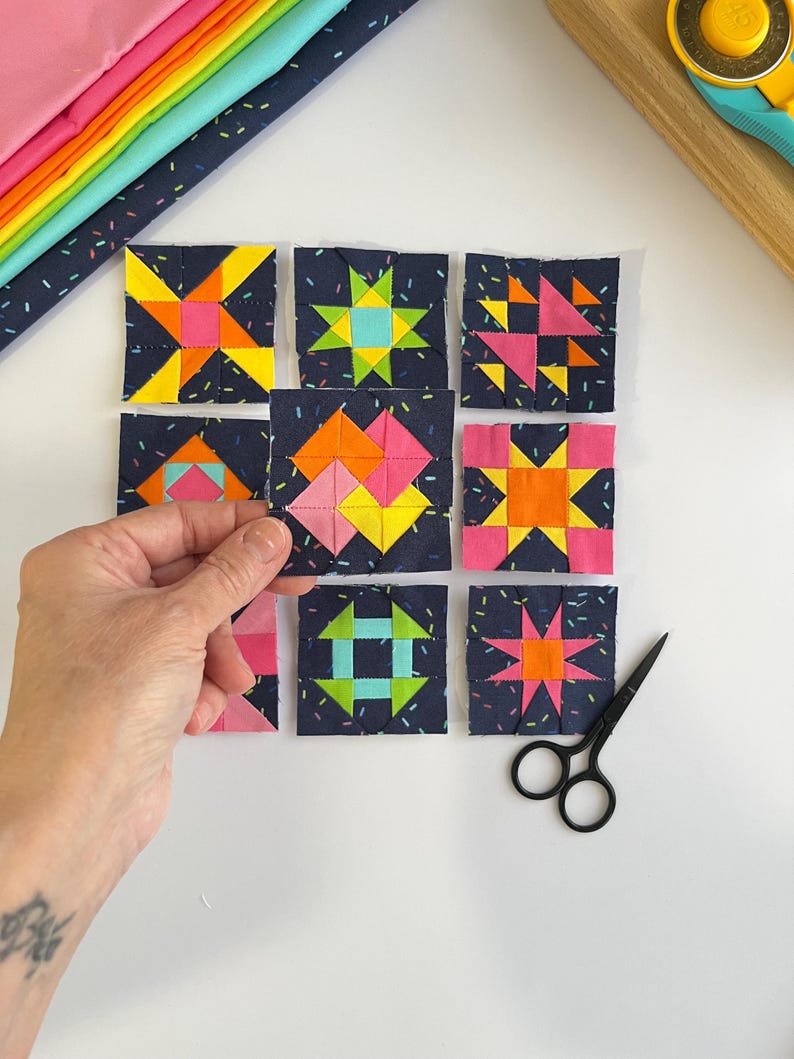 Peut inclure: Une collection de neuf blocs de courtepointe en tissu carr&eacute; dispos&eacute;s en grille. Chaque bloc pr&eacute;sente un fond bleu marine fonc&eacute; avec un imprim&eacute; confettis et des motifs g&eacute;om&eacute;triques en tissus jaune vif, orange, rose et bleu clair.