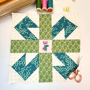 Pode incluir: Um bloco de colcha de retalhos com um padrão verde, branco e azul. O centro do bloco apresenta uma pequena ilustração de um rato rosa segurando um molho de cenouras.