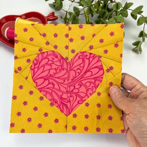 Pode incluir: Um bloco de tecido quadrado com um desenho de coração rosa sobre um fundo amarelo. O coração é preenchido com um padrão de redemoinho. O tecido amarelo tem um pequeno padrão floral rosa. O bloco provavelmente faz parte de um edredom ou outro projeto têxtil.