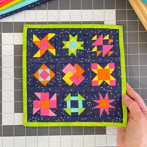 Peut inclure: Un bloc de courtepointe en tissu carr&eacute; pr&eacute;sentant neuf motifs g&eacute;om&eacute;triques distincts dans des couleurs n&eacute;on vives, notamment rose, orange, jaune et bleu, sur un fond bleu marine fonc&eacute; avec un imprim&eacute; confettis et une bordure en tissu vert lime.