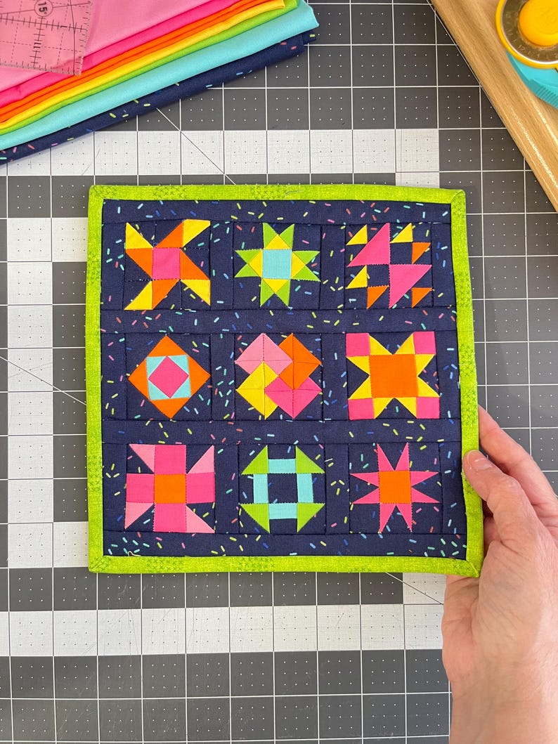 Peut inclure: Un bloc de courtepointe en tissu carr&eacute; pr&eacute;sentant neuf motifs g&eacute;om&eacute;triques distincts dans des couleurs n&eacute;on vives, notamment rose, orange, jaune et bleu, sur un fond bleu marine fonc&eacute; avec un imprim&eacute; confettis et une bordure en tissu vert lime.