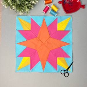 Pode incluir: Um bloco de colcha geométrico colorido com um design de estrela. A estrela é composta por triângulos laranja, rosa e amarelo sobre um fundo azul claro. Materiais de costura, incluindo carretéis de linha e tesouras, são dispostos ao redor do bloco.