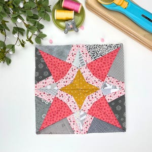 Pode incluir: Um bloco de colcha quadrado com um design de estrela. O bloco de colcha apresenta vários tecidos em tons de rosa, cinza e amarelo. O centro da estrela é amarelo e as pontas são rosa. O bloco é provavelmente para um projeto de colcha maior.