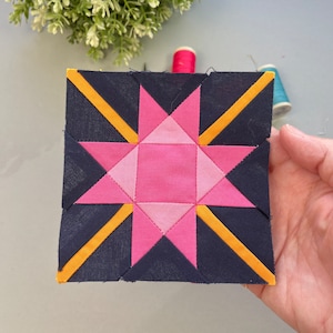 Pode incluir: Um bloco de tecido quadrado para colcha com um desenho de estrela. O bloco tem um fundo azul marinho com uma estrela rosa no centro, rodeada por triângulos rosa mais claros. Listras diagonais amarelas adicionam contraste. Materiais de costura no fundo.