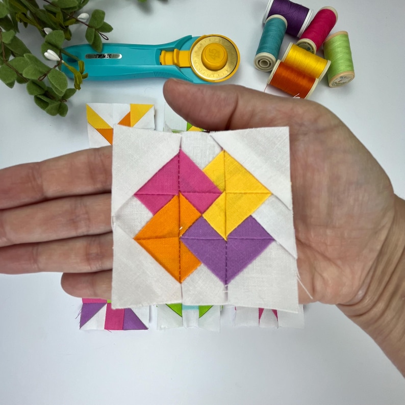 Peut inclure: Un bloc de courtepointe carr&eacute; en tissu pr&eacute;sentant un motif central de moulin &agrave; vent compos&eacute; de triangles roses, jaunes, orange et violets sur un fond blanc. Le bloc est tenu, avec un cutter rotatif turquoise et des bobines de fil color&eacute;es.