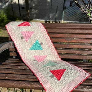 May include: A white table runner with a red, pink and teal geometric pattern of Christmas trees. The runner is on a wooden bench.