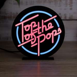 May include: A black circular neon sign with a blue and pink neon outline. The sign says "Top of the Pops" in pink neon lettering.