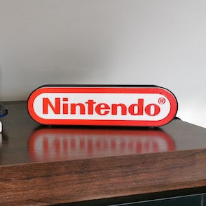 May include: A black Nintendo 3DS handheld gaming console with a blue screen and a game cartridge inserted. The console is sitting on top of a game case. A red and white Nintendo logo is on a black rectangular speaker.