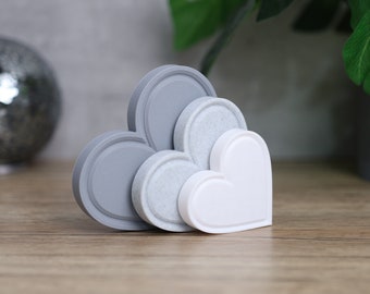 May include: A collection of heart-shaped and circular decorative objects in shades of gray and white. The objects are arranged together on a wooden surface, with a decorative sphere and greenery in the background.