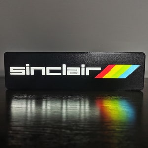May include: A black rectangular sign with the word "Sinclair" in white letters and a rainbow stripe design.
