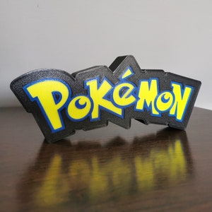 May include: A black 3D printed sign with the word "Pokemon" in yellow letters with blue outlines.