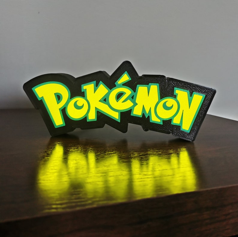 May include: A black and green 3D printed sign with the word "Pok&eacute;mon" in yellow letters.