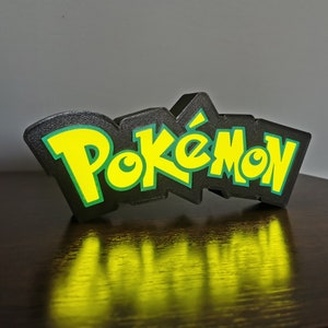 May include: Black and green 3D printed Pok&eacute;mon logo. The logo is made of plastic and has a glossy finish.