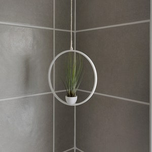 May include: A white metal hanging planter with a green air plant inside. The planter is hanging from a white string against a white tiled wall.