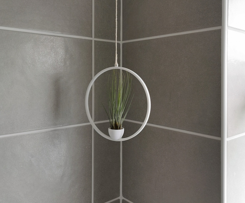 May include: A white metal hanging planter with a green air plant inside. The planter is hanging from a white string against a white tiled wall.