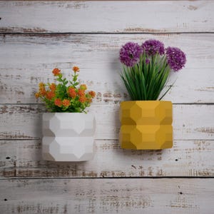 May include: Two geometric planters with different coloured flowers. The planter on the left is white and the planter on the right is yellow. The flowers in the white planter are orange and the flowers in the yellow planter are purple.