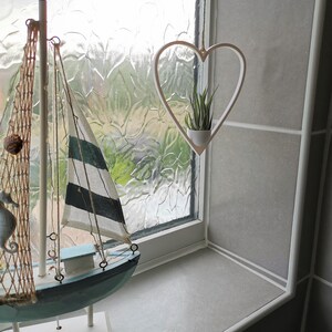 May include: A white heart-shaped hanging planter with a small green plant inside. The planter is hanging from a window with a white frame. A decorative wooden sailboat is in the foreground.