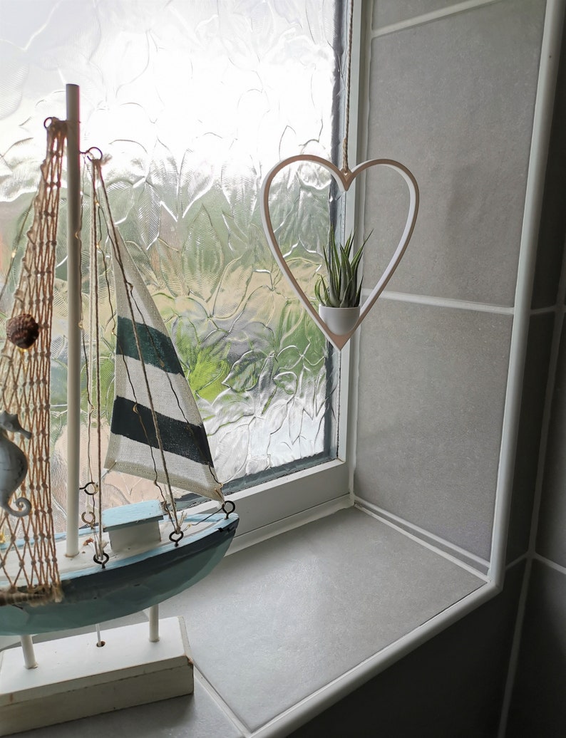 May include: A white heart-shaped hanging planter with a small green plant inside. The planter is hanging from a window with a white frame. A decorative wooden sailboat is in the foreground.