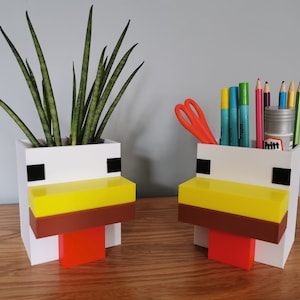 May include: Two white ceramic chicken-shaped desk organisers. One organiser holds a potted plant with green leaves. The other organiser holds pens, pencils, and a pair of scissors.