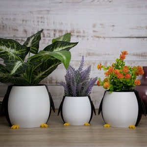 May include: Three white penguin-shaped planters with black accents. The planters are filled with green, purple, and orange plants. The planters are sitting on a wooden surface.
