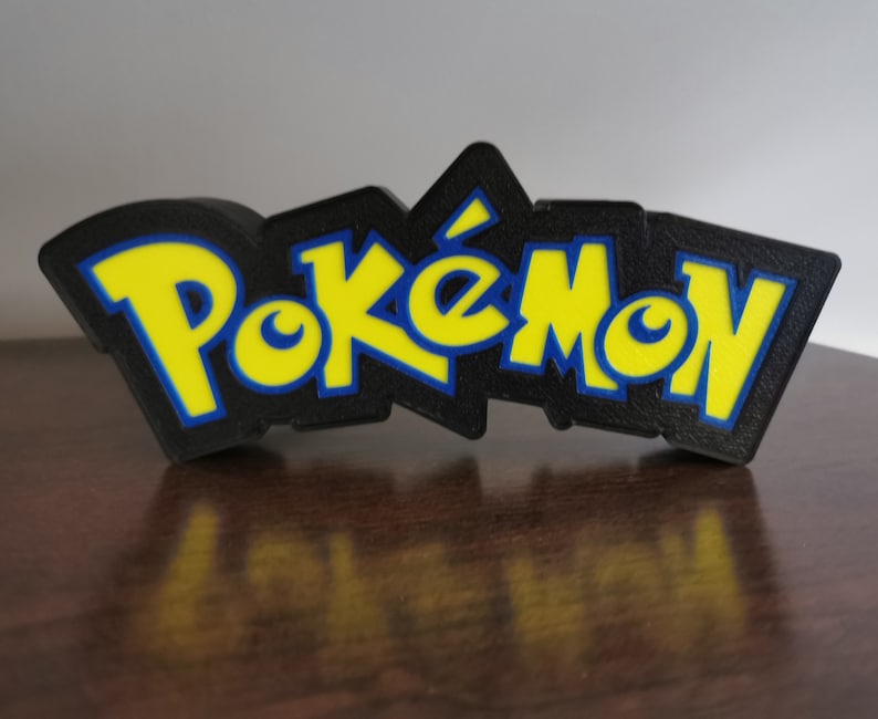 May include: A 3D printed black, yellow, and blue Pok&eacute;mon logo. The logo is raised and has a textured surface.