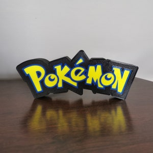May include: A black 3D printed sign with the word "Pok&eacute;mon" in yellow letters with blue outlines.