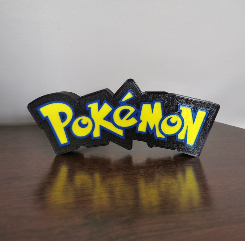 May include: A black 3D printed sign with the word "Pok&eacute;mon" in yellow letters with blue outlines.