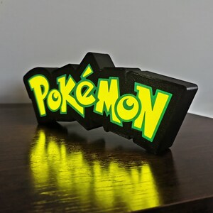 May include: A black and green 3D printed sign with the word "Pok&eacute;mon" in yellow letters.