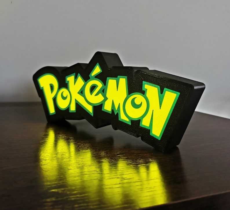 May include: A black and green 3D printed sign with the word "Pok&eacute;mon" in yellow letters.