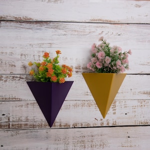 May include: Two triangular wall planters, one purple and one yellow, with artificial flowers. The purple planter has orange flowers and the yellow planter has pink flowers.