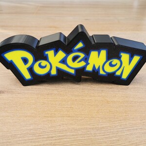 May include: A black 3D printed Pokemon logo with yellow letters. The logo is on a wooden surface.