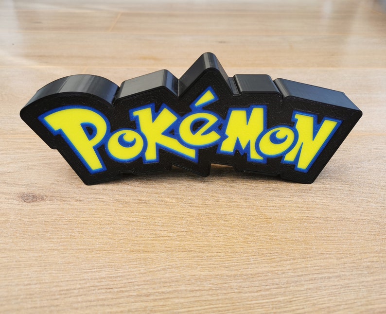 May include: A black 3D printed Pokemon logo with yellow letters. The logo is on a wooden surface.