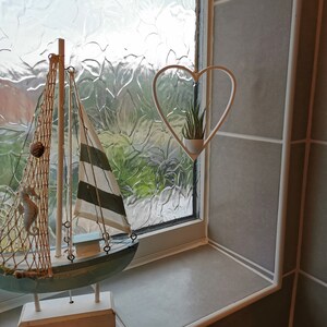 May include: A white wooden sailboat with a blue hull and a white and green striped sail. The sailboat is on a white stand and is in front of a window with a white heart-shaped planter hanging from the top. The planter has a small green plant inside.