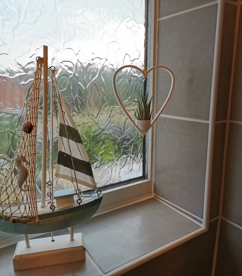 May include: A white wooden sailboat with a blue hull and a white and green striped sail. The sailboat is on a white stand and is in front of a window with a white heart-shaped planter hanging from the top. The planter has a small green plant inside.