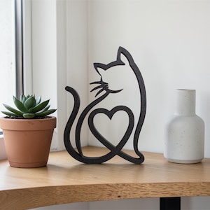 May include: A black metal cat silhouette with a heart-shaped design. The cat sculpture sits on a wooden surface next to a small potted succulent and a white ceramic vase. The cat's tail curves into a heart.