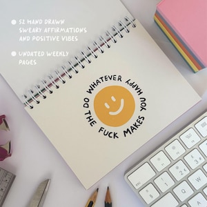 May include: A spiral-bound notebook with the text "WHATEVER YOU HAPPY MAKES DO THE FUCK" around a yellow smiley face. The notebook also has the text "52 HAND DRAWN SWEARY AFFIRMATIONS AND POSITIVE VIBES" and "UNDATED WEEKLY PAGES". A keyboard and sticky notes are also visible.