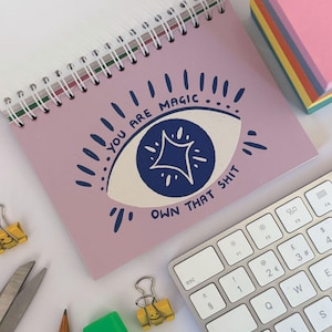 May include: A spiral-bound notebook with a pink cover featuring a blue and white eye design and the text "YOU ARE MAGIC OWN THAT SHIT." A keyboard, sticky notes, and office supplies are also visible.