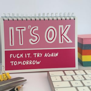 May include: A desk calendar with a pink and magenta color scheme. The text "IT'S OK" is displayed in large white letters. Below, the text reads "FUCK IT. TRY AGAIN TOMORROW". A keyboard, scissors, and sticky notes are also visible.