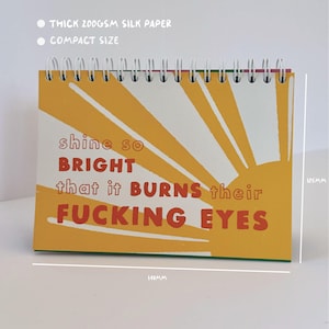 May include: A compact, spiral-bound notepad with a yellow sunburst design and the text "shine so bright that it burns their fucking eyes" in red. The notepad is 148mm wide and 105mm tall, made with thick 200gsm silk paper.