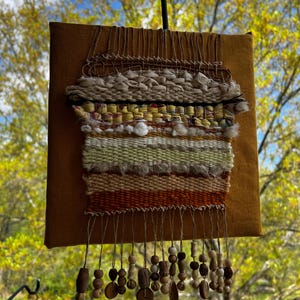 May include: A woven wall hanging with a square, mustard-colored backing. The woven portion features a mix of textures and colors, including cream, yellow, and orange. Wooden beads and shapes dangle from the bottom, adding a decorative element. The piece is displayed outdoors.