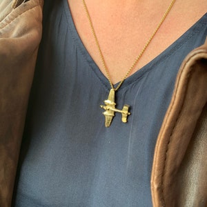 May include: A gold chain necklace with a gold airplane pendant. The necklace is worn by a person wearing a blue shirt and a brown leather jacket.