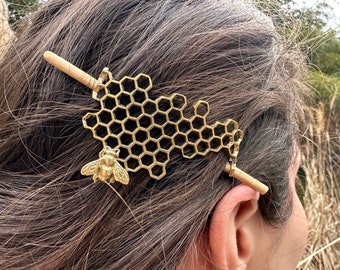 May include: A gold-toned hair stick with a honeycomb design and a bee charm. The stick is made of wood and has a natural finish.