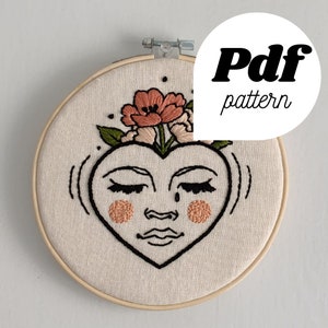 Peut inclure: Art de broderie sur un cercle en bois naturel représentant un visage en forme de cœur avec les yeux fermés et une seule larme, orné d'une couronne florale de fleurs roses et vertes.