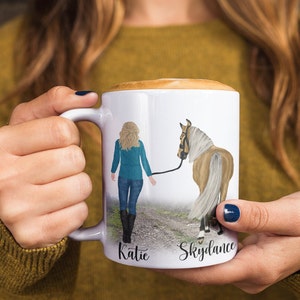May include: White ceramic mug with a colorful illustration of a woman walking a horse. The mug has the names "Katie" and "Skydance" written on the bottom.