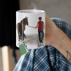 May include: White ceramic mug with a brown horse and a person in a red jacket walking on a path. The mug has the names "Dakota" and "Tyler" written on it.