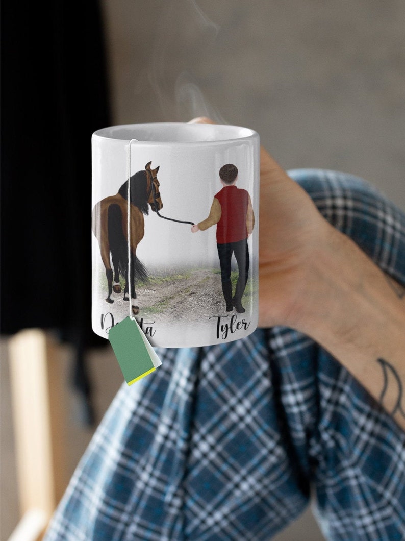 May include: White ceramic mug with a brown horse and a person in a red jacket walking on a path. The mug has the names "Dakota" and "Tyler" written on it.